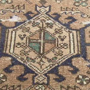 Peut inclure: Gros plan sur un tapis vintage avec un motif g&eacute;om&eacute;trique central. Le tapis pr&eacute;sente une palette de couleurs chaudes de marron, beige et bleu marine. Le motif comprend une forme de losange central avec des d&eacute;tails complexes et des motifs floraux.