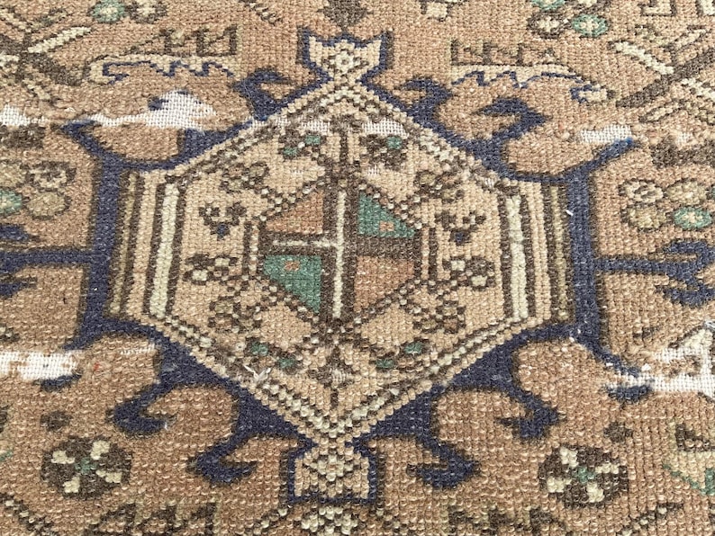 Peut inclure: Gros plan sur un tapis vintage avec un motif g&eacute;om&eacute;trique central. Le tapis pr&eacute;sente une palette de couleurs chaudes de marron, beige et bleu marine. Le motif comprend une forme de losange central avec des d&eacute;tails complexes et des motifs floraux.