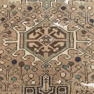 Peut inclure: Gros plan sur un tapis vintage avec un motif g&eacute;om&eacute;trique d&eacute;taill&eacute;. Le tapis pr&eacute;sente un m&eacute;daillon central dans des tons de marron, beige et sarcelle, entour&eacute; de motifs floraux et abstraits complexes. La bordure du tapis est marron fonc&eacute; avec des motifs r&eacute;p&eacute;titifs.