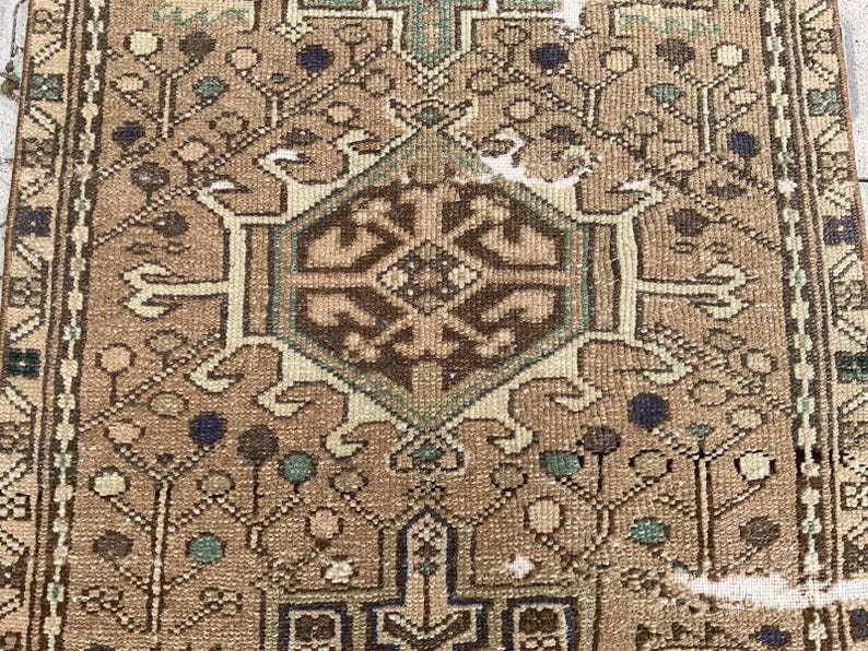 Peut inclure: Gros plan sur un tapis vintage avec un motif g&eacute;om&eacute;trique d&eacute;taill&eacute;. Le tapis pr&eacute;sente un m&eacute;daillon central dans des tons de marron, beige et sarcelle, entour&eacute; de motifs floraux et abstraits complexes. La bordure du tapis est marron fonc&eacute; avec des motifs r&eacute;p&eacute;titifs.