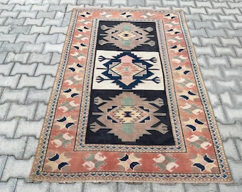 Könnte beinhalten: Ein rechteckiger Teppich mit einem zentralen schwarzen Feld, das drei geometrische Medaillons in Rosa-, Beige- und Dunkelblautönen aufweist. Der Teppich ist von einer breiten terrakottafarbenen Bordüre mit einem wiederkehrenden Muster kleiner Motive umrahmt.