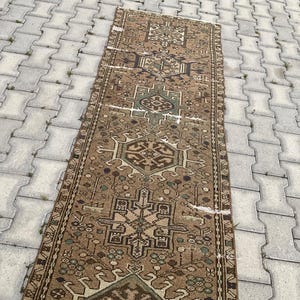 Peut inclure: Un tapis rectangulaire de style vintage avec un motif g&eacute;om&eacute;trique d&eacute;taill&eacute; dans des tons de marron, beige et vert. Le tapis pr&eacute;sente un motif central avec des motifs r&eacute;p&eacute;titifs et une bordure. Le tapis est pos&eacute; sur une surface grise textur&eacute;e.
