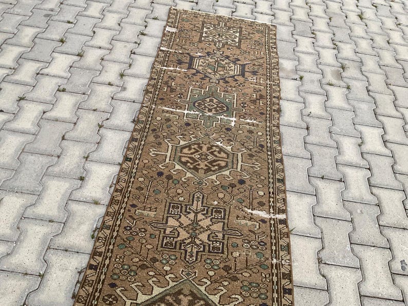 Peut inclure: Un tapis rectangulaire de style vintage avec un motif g&eacute;om&eacute;trique d&eacute;taill&eacute; dans des tons de marron, beige et vert. Le tapis pr&eacute;sente un motif central avec des motifs r&eacute;p&eacute;titifs et une bordure. Le tapis est pos&eacute; sur une surface grise textur&eacute;e.