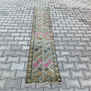 May include: A rectangular, vintage-style runner rug with a floral pattern in shades of pink, brown, and green. The rug has a dark green border and is placed on a gray brick surface. The rug's design features repeating floral motifs.