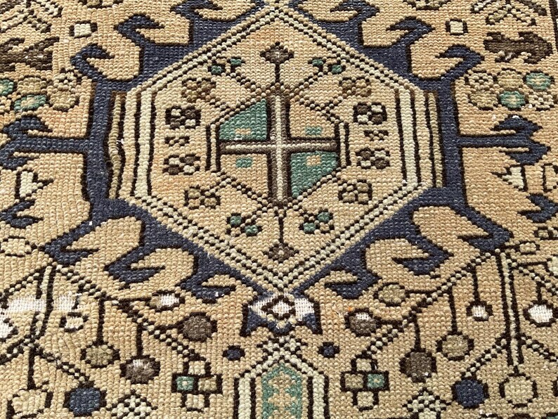 Peut inclure: Gros plan sur un tapis beige et bleu marine &agrave; motifs. Le tapis pr&eacute;sente une forme de losange central avec un motif de croix au centre, entour&eacute; de motifs floraux et g&eacute;om&eacute;triques complexes. Le tapis a un style vintage et traditionnel.