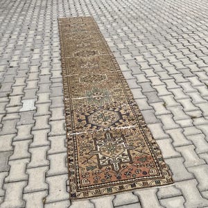 Peut inclure: Un long tapis rectangulaire vintage avec un motif g&eacute;om&eacute;trique d&eacute;taill&eacute; dans les tons de marron, beige, vert et rouge. Le tapis est pos&eacute; sur une surface en briques grises, mettant en valeur son design complexe et son aspect vieilli. Les dimensions du tapis ne sont pas visibles.