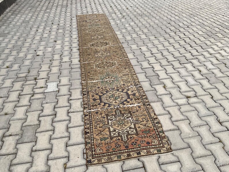 Peut inclure: Un long tapis rectangulaire vintage avec un motif g&eacute;om&eacute;trique d&eacute;taill&eacute; dans les tons de marron, beige, vert et rouge. Le tapis est pos&eacute; sur une surface en briques grises, mettant en valeur son design complexe et son aspect vieilli. Les dimensions du tapis ne sont pas visibles.