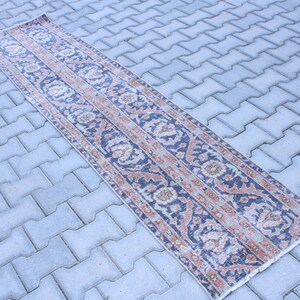 Könnte beinhalten: Ein langer, schmaler Teppich mit einem blauen und rosafarbenen Blumenmuster. Der Teppich hat einen verblassten, Vintage-Look und liegt auf einer grauen Betonfläche.