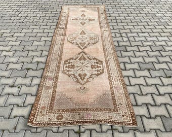 Op de afbeelding: Een rechthoekig vloerkleed in vintage stijl met een vervaagde uitstraling. Het vloerkleed heeft een centraal ontwerp in bruin- en beigetinten, met geometrische patronen en bloemaccenten. De rand van het vloerkleed heeft een gedetailleerd, sierlijk ontwerp.