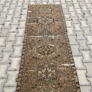 Peut inclure: Un tapis rectangulaire de style vintage avec un motif g&eacute;om&eacute;trique d&eacute;taill&eacute;. Le tapis pr&eacute;sente un motif central dans des tons de marron, beige et vert, avec des bordures complexes. Le tapis est pos&eacute; sur une surface grise textur&eacute;e.