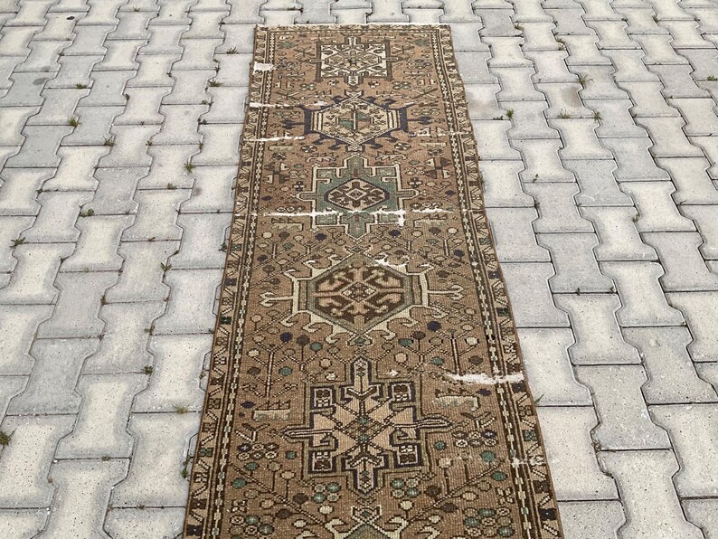 Peut inclure: Un tapis rectangulaire de style vintage avec un motif g&eacute;om&eacute;trique d&eacute;taill&eacute;. Le tapis pr&eacute;sente un motif central dans des tons de marron, beige et vert, avec des bordures complexes. Le tapis est pos&eacute; sur une surface grise textur&eacute;e.