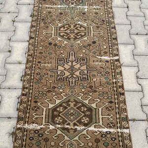 Peut inclure: Un tapis rectangulaire de style vintage avec un motif g&eacute;om&eacute;trique central dans des tons de marron, beige et vert. Le tapis pr&eacute;sente des motifs complexes et un aspect vieilli, sugg&eacute;rant l'&acirc;ge et l'utilisation. Le tapis est pos&eacute; sur une surface grise textur&eacute;e.
