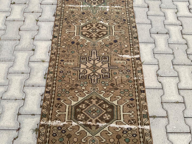 Peut inclure: Un tapis rectangulaire de style vintage avec un motif g&eacute;om&eacute;trique central dans des tons de marron, beige et vert. Le tapis pr&eacute;sente des motifs complexes et un aspect vieilli, sugg&eacute;rant l'&acirc;ge et l'utilisation. Le tapis est pos&eacute; sur une surface grise textur&eacute;e.