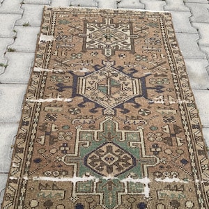 Peut inclure: Un tapis rectangulaire vintage avec un motif g&eacute;om&eacute;trique d&eacute;taill&eacute;. Le tapis pr&eacute;sente un motif central dans des tons de marron, de bleu et de vert, avec des bordures complexes. L'aspect vieilli du tapis ajoute &agrave; son caract&egrave;re.
