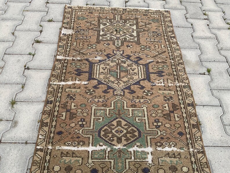 Peut inclure: Un tapis rectangulaire vintage avec un motif g&eacute;om&eacute;trique d&eacute;taill&eacute;. Le tapis pr&eacute;sente un motif central dans des tons de marron, de bleu et de vert, avec des bordures complexes. L'aspect vieilli du tapis ajoute &agrave; son caract&egrave;re.