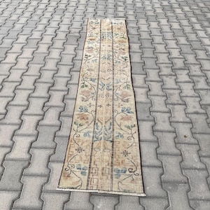May include: A rectangular, beige runner rug with a floral pattern in shades of blue, brown, and green. The rug is placed on a gray, patterned surface. The rug's design features vertical stripes with floral and leaf motifs.