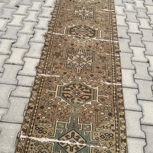 Peut inclure: Un tapis rectangulaire de style vintage avec un motif g&eacute;om&eacute;trique complexe dans des tons de marron, de vert et de beige. Le tapis est pos&eacute; sur une surface grise et textur&eacute;e. Le design du tapis pr&eacute;sente des motifs et des bordures r&eacute;p&eacute;titifs.
