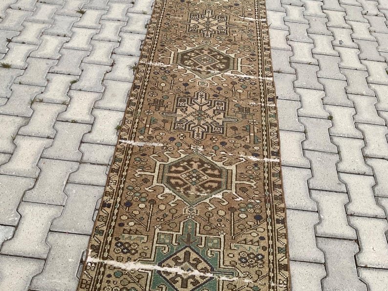 Peut inclure: Un tapis rectangulaire de style vintage avec un motif g&eacute;om&eacute;trique complexe dans des tons de marron, de vert et de beige. Le tapis est pos&eacute; sur une surface grise et textur&eacute;e. Le design du tapis pr&eacute;sente des motifs et des bordures r&eacute;p&eacute;titifs.