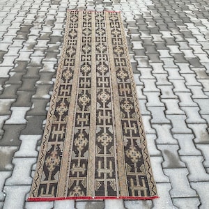 Pode incluir: Um tapete retangular com um padrão geométrico castanho e bege. O tapete tem uma borda vermelha e está colocado sobre uma superfície cinzenta texturizada. O design apresenta listras verticais com formas repetidas.