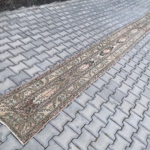 May include: A long, narrow, rectangular rug with an intricate, faded floral pattern in shades of brown, beige, and pink. The rug is placed on a gray, patterned surface, showcasing its length and detailed design. The rug's edges are finished with a white border.