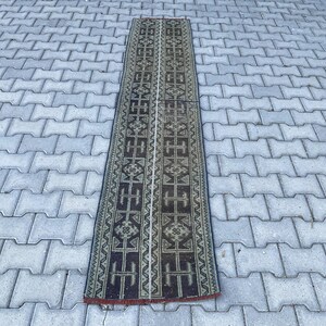 May include: A long, narrow runner rug featuring a dark brown and muted olive green geometric pattern. The design consists of repeating symmetrical motifs, including stylised cross shapes and intricate borders, arranged in a vertical orientation.