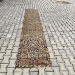 Peut inclure: Un long tapis rectangulaire vintage avec un motif g&eacute;om&eacute;trique complexe. Le tapis pr&eacute;sente un motif central dans des tons de marron, vert et beige, avec une bordure plus fonc&eacute;e. Le tapis est pos&eacute; sur une surface en briques grises.