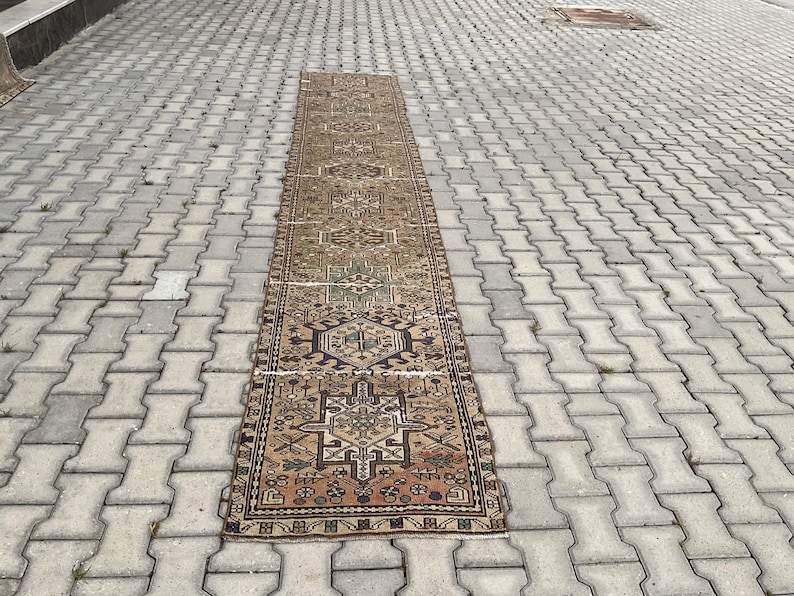 Peut inclure: Un long tapis rectangulaire vintage avec un motif g&eacute;om&eacute;trique complexe. Le tapis pr&eacute;sente un motif central dans des tons de marron, vert et beige, avec une bordure plus fonc&eacute;e. Le tapis est pos&eacute; sur une surface en briques grises.