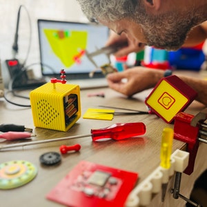 May include: A yellow and red electronic device with a speaker grille and a red button on the top. The device is sitting on a wooden table with other electronic components and tools.