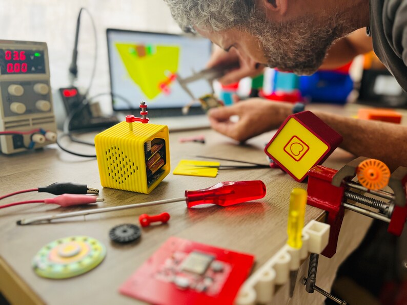 May include: A yellow and red electronic device with a speaker grille and a red button on the top. The device is sitting on a wooden table with other electronic components and tools.