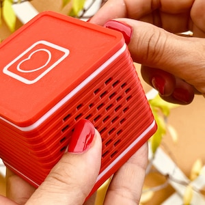 May include: A red and white portable speaker with a heart-shaped icon on the front. The speaker has a grid pattern on the side.
