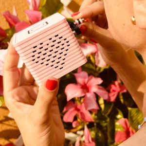 May include: A pink speaker with a heart-shaped speaker grille. The speaker is being held by a person's hand with pink nail polish.