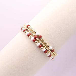 May include: Three beaded bracelets with gold, red, and white beads. The bracelets are stacked together, showcasing the different colors and textures. The gold beads provide a metallic accent, while the red and white beads add pops of color.