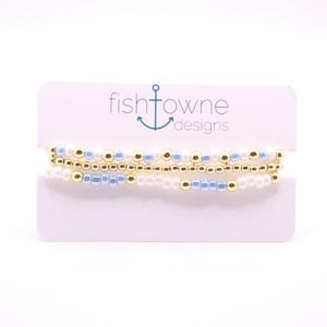 May include: Three beaded bracelets in gold, white, and light blue are displayed on a white card. The bracelets are made of small, round beads. The card has the text "fishtowne designs" with an anchor logo.