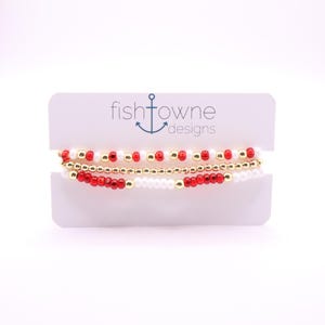 May include: Three beaded bracelets in red, white, and gold are displayed on a white card. The top bracelet has red and gold beads, the middle one is gold, and the bottom one has red and white beads. The card reads "fishtowne designs".
