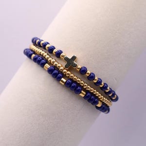 May include: Three beaded bracelets. The bracelets feature navy blue and gold beads. One bracelet has a small gold cross charm. The bracelets are stacked on top of each other.