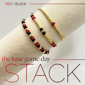 May include: Three beaded bracelets in red, black, and gold are displayed. The bracelets are stacked on a white surface. The text "RED + BLACK" and "the luxe game day STACK" are visible in the image.