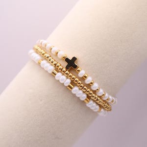 May include: Stacked beaded bracelets in gold, white, and black. The bracelets feature small gold beads, white beads, and a black cross charm. The bracelets are stacked together, creating a layered look.