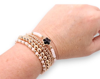 May include: A stack of gold beaded bracelets with white and gold accents. One bracelet features a black clover charm. The bracelets are worn on a wrist against a white background.