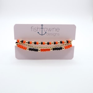May include: Three beaded bracelets in orange, black, and gold are displayed on a white card. The bracelets are made of small, round beads. The card has the text "fishtowne designs" with an anchor logo.
