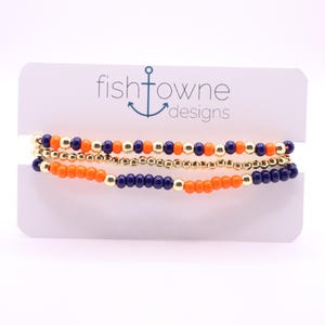 May include: Three beaded bracelets in orange, navy blue, and gold. The bracelets are displayed on a white card with the text "fishtowne designs" and an anchor logo. The beads are small and evenly spaced.