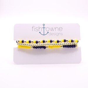 May include: Three beaded bracelets in yellow, navy blue, and gold. The bracelets are displayed on a white card with the text "fishtowne designs" and an anchor logo. The bracelets are made of small, round beads.