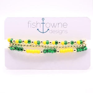 May include: Three beaded bracelets in yellow, green, and gold. The bracelets are displayed on a white card with the text "fishtowne designs" and an anchor logo. The beads are small and evenly spaced, creating a delicate and colorful accessory.