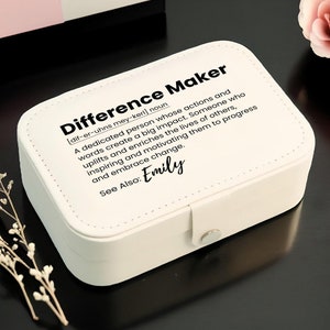 May include: White jewelry box with a black inscription that reads "Difference Maker [dif-er-uhns mey-ker] noun A dedicated person whose actions and words create a big impact. Someone who uplifts and enriches the lives of others, inspiring and motivating them to progress and embrace change. See Also: Emily"