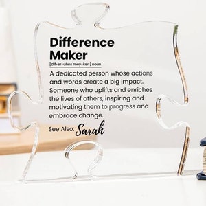 May include: A clear acrylic puzzle piece with the word "Difference Maker" defined as "a dedicated person whose actions and words create a big impact." The text also says "See Also: Sarah".