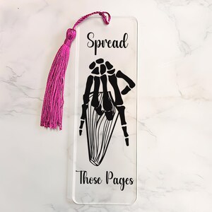 May include: A clear acrylic bookmark with a black skeleton hand design and the text "Spread Those Pages". The bookmark has a pink tassel.