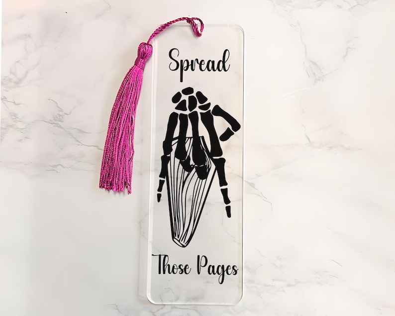 May include: A clear acrylic bookmark with a black skeleton hand design and the text "Spread Those Pages". The bookmark has a pink tassel.
