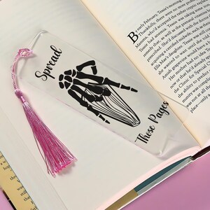 May include: A clear acrylic bookmark with a pink tassel. The bookmark features a black silhouette of a skeleton hand making the "spread" hand gesture. The text "Spread" is printed in black at the top of the bookmark. The text "Those Pages" is printed in black at the bottom of the bookmark.