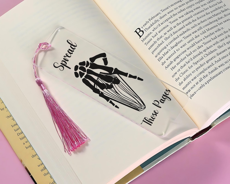 May include: A clear acrylic bookmark with a pink tassel. The bookmark features a black silhouette of a skeleton hand making the "spread" hand gesture. The text "Spread" is printed in black at the top of the bookmark. The text "Those Pages" is printed in black at the bottom of the bookmark.