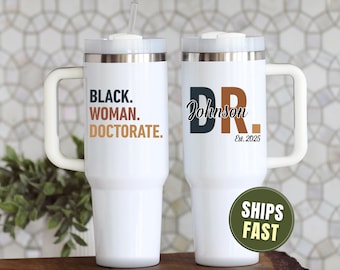 May include: Two white insulated travel mugs with handles. One mug has the text "BLACK. WOMAN. DOCTORATE." in black, brown, and orange. The other mug has "DR. Johnson Est. 2025" in black and brown. Both mugs have a white lid and a straw.