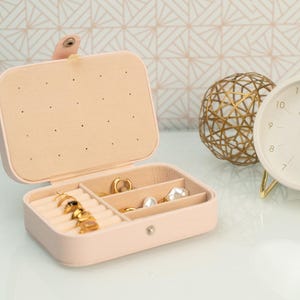 May include: A pink faux leather jewelry box with a hinged lid and multiple compartments for storing rings, earrings, and other small jewelry items. The box has a gold-colored clasp and is open to reveal the compartments.